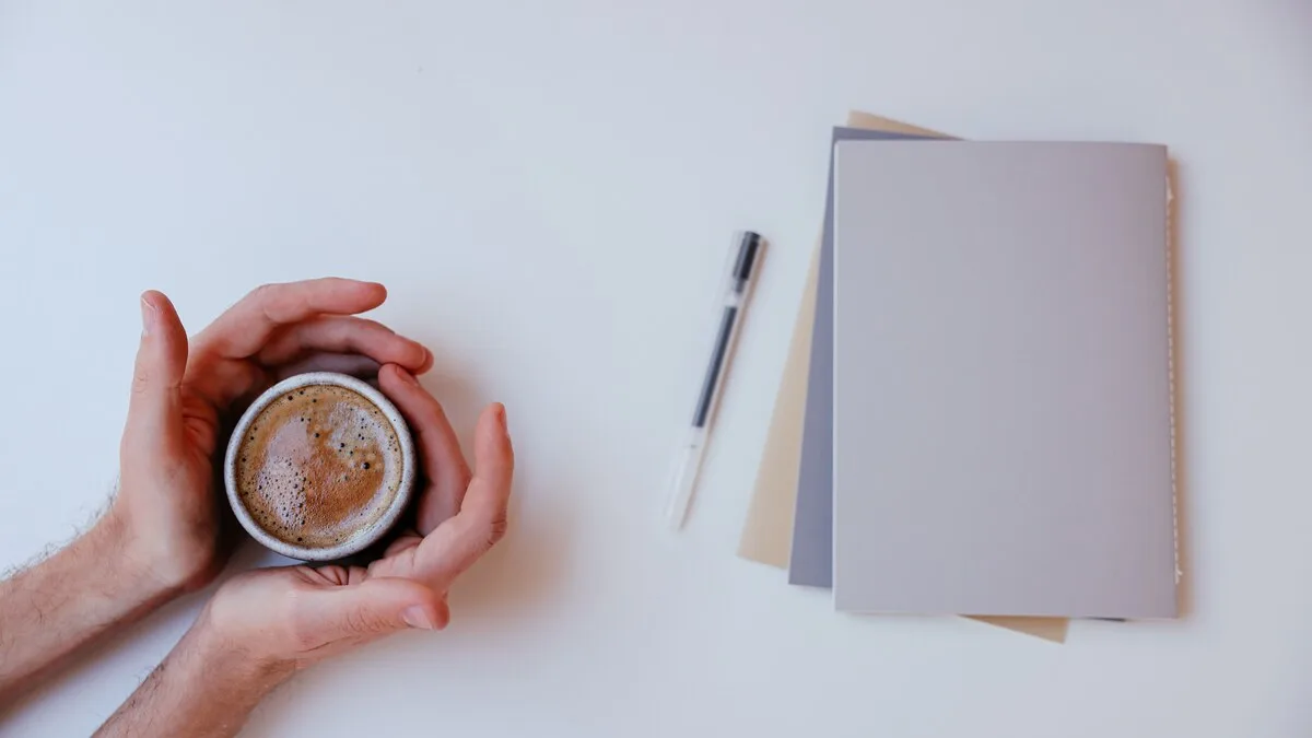 A pair of hands cradling a coffee cup beside two notebooks and a pen
